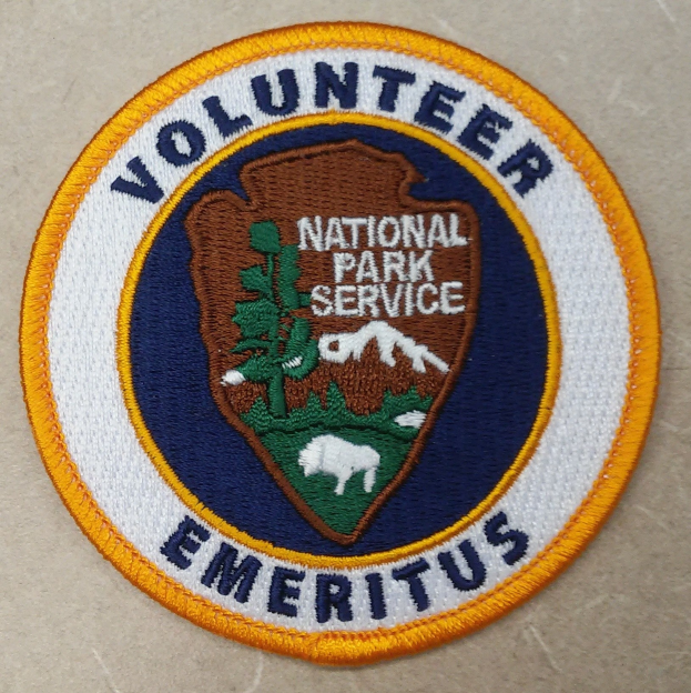 Rundes Abzeichen mit blauem Rand und wei├čem Hintergrund, das die zentrierte schwarze Schrift "Volunteer National Park Service Emeritus" in einem gelben Kreis zeigt.