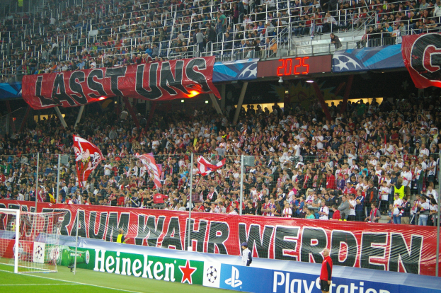 Fußballspiel in einem Stadion mit großer Menschenmenge, saftigem Grün, Torpfosten links, Bodenbanner, Menschen mit Fahnen und Hintergrundgeländer, Tafeln und Lichter.