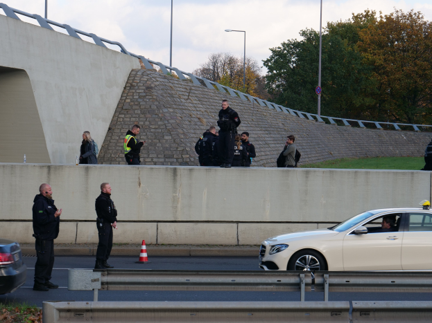 Eine Gruppe von Polizeibeamten steht neben einem Auto auf der Straße, mit Verkehrskegeln, einer Barriere, Gras, einer Wand, Laternenpfählen, Bäumen und einem bewölkten Himmel im Hintergrund.
