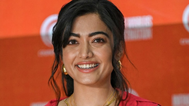 Eine Frau in einem roten Kleid mit einem eleganten Dutt, schwarzen High Heels und natürlichem Make-up lächelt in die Kamera, mit einem gelben Banner im Hintergrund, das 'Die besten Schönheits Looks von Sonam Kapoor beim Cannes Film Festival' trägt.