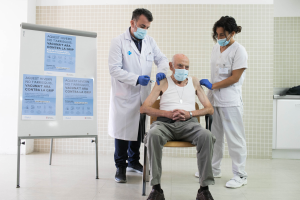 Ein älterer Mann sitzt in einem Stuhl und erhält eine Impfung von zwei maskierten und handschuhgetragenen Ärzten in einer Krankenhausumgebung, mit einer Texttafel links und Gegenständen auf einer Plattform rechts.