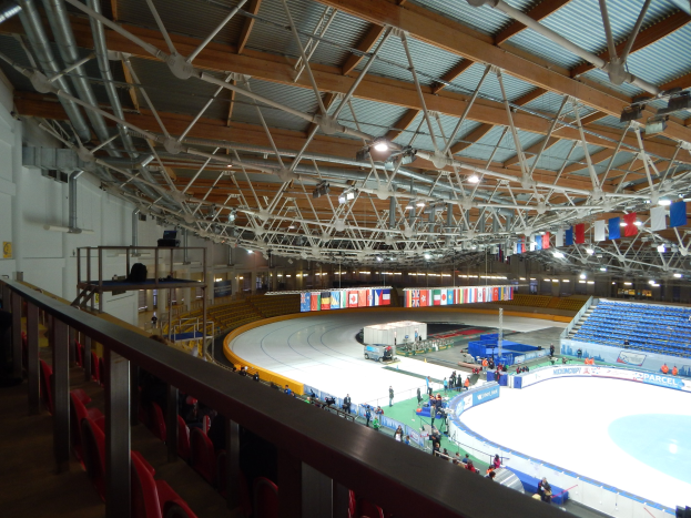 Großes Indoor-Eisstadion mit zahlreichen Menschen auf dem Eis, umgeben von Sitzplätzen, Geländern, Brettern, Beleuchtungskörpern und konstruktiven Elementen, mit einer Wand im Hintergrund.