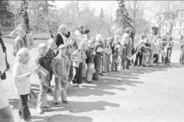 Schwarz-weißes Bild einer Protestdemonstration auf einem Schulgelände, mit Menschen, die Fahnen in einer Reihe auf einem Schotterweg halten, Bäume und Gebäude im Hintergrund.