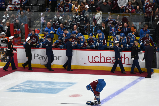 Ein Hockey-Spieler in dunkler Uniform hält einen Schläger auf dem Eis, umgeben von Menschen in Mützen und mit Hockey-Schlägern, mit Zuschauern auf Sitzen und Stehplätzen im Hintergrund hinter Brettern, die "Colorado Avalanche vs San Jose Sharks - 3/24/15 NHL Pick, Odds, and Prediction" anzeigen.