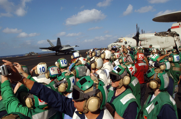 Eine Gruppe von Menschen in Helmen und Schutzbrillen auf einem Flugzeugträger, mit einem Flugzeug im Hintergrund und Wolken am Himmel, einige halten Kameras.