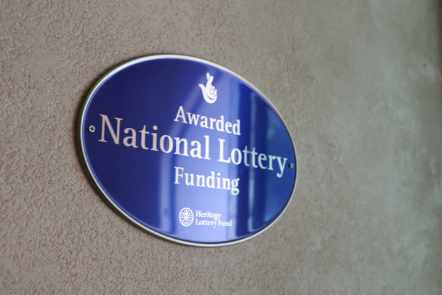 Blaues rechteckiges Schild an einer Gebäudewand mit weißem Hintergrund und schwarzer fetter Schrift, die "National Lottery Funding awarded" liest.