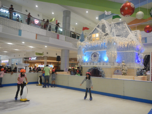 Eine Gruppe von Menschen, die auf einer Eisbahn in einem Einkaufszentrum Schlittschuh laufen, umgeben von einem hausähnlichen Aufbau, der mit Lichtern und festlichen Dekorationen geschmückt ist, mit Menschen in der Nähe und Lichtern an der Decke, die eine festliche Atmosphäre schaffen.