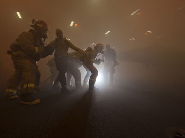 Feuerwehrleute in Helmen gehen öffentlich eine neblige Straße entlang und halten Gegenstände, mit einem Flugzeug und Lichtern im Hintergrund und einer Straße unten.