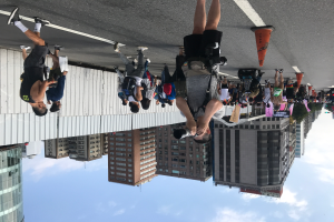 Eine Gruppe von Menschen steht an der Straße, ein Mann hält eine Kamera in den Vordergrund, Gebäude mit Fenstern, Bäume und ein bewölkter Himmel im Hintergrund und ein Verkehrskegel auf der rechten Seite.