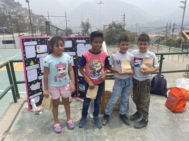 Gruppe von Kindern auf einem Basketballfeld mit Papiertüten in den Händen, mit Tüten und Gegenständen rechts und einer Tafel mit Text links, vor dem Hintergrund von Zaun, Pfosten, Straßenlaternen, Bäumen, Gebäuden, Hügeln und Himmel.