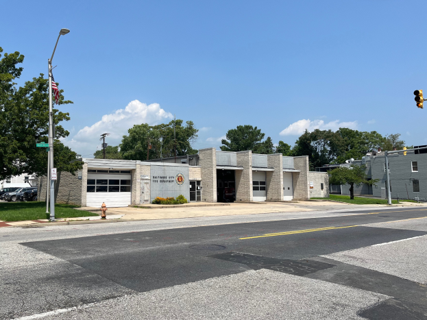 Eine Feuerwache an einer Straßenecke mit umliegenden Gebäuden, Fahrzeugen, Verkehrszeichen, einer Feuerlöschstelle, Vegetation und einem bewölkten Himmel.