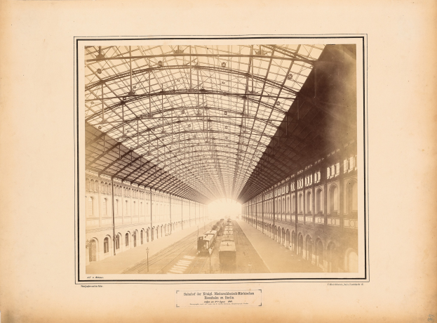 Schwarz-weiß-Foto eines Bahnhofs mit einem Zug auf den Schienen, umgeben von Gebäuden mit Fenstern und einem Dach, das von Metallpfosten gestützt wird, mit Text unten.