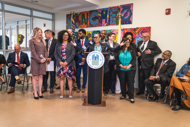 Eine Gruppe von Menschen, die vor einem Rednerpult mit Mikrofon und einer Tafel mit Text darauf stehen und sitzen. Im Hintergrund sind Flaggen, eine Tafel, ein Feuerlöscher und Deckenleuchten zu sehen.