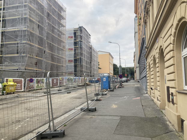 Stadtstraße mit Gebäuden, Fahrzeugen, Straßeninfrastruktur, Bäumen und einer Baustelle mit Gerüsten im Vordergrund.
