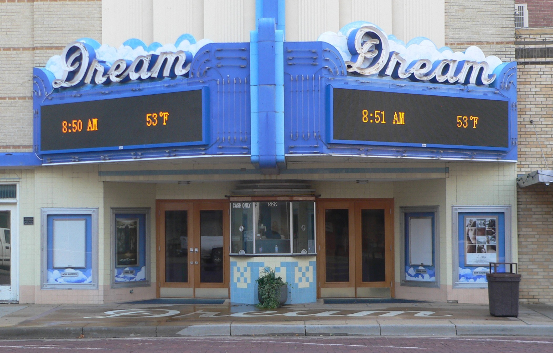Außenansicht des Dream Theaters in St. Louis, Missouri mit Glasfenstern und -türen, einem Schild, einem Mülleimer, einem Topf mit einer Pflanze und einer Straße unten.