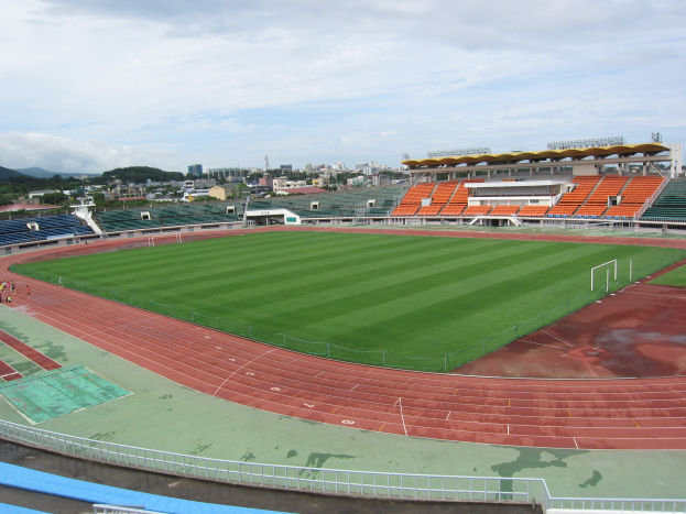 Großes Stadion mit einem Fußballfeld, umgeben von Gebäuden, Bäumen, Hügeln und einem klaren blauen Himmel, mit saftigem Grün und wenigen Menschen.
