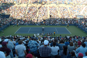 Eine große Menschenmenge schaut einem Tennisspiel in einem ausverkauften Stadion zu, mit Spielern auf dem Platz und Zuschauern in den Rängen.