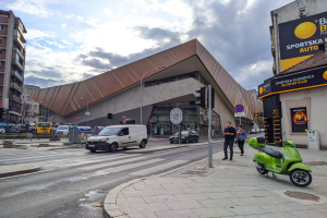 Ein grüner E-Scooter steht auf der Seite einer Straße neben einem Gebäude, mit Fahrzeugen auf der Straße, Fußgängern auf dem Gehweg und Verkehrsampeln mit Masten, während Gebäude mit Fenstern, Bäume und ein bewölkter Himmel im Hintergrund zu sehen sind.