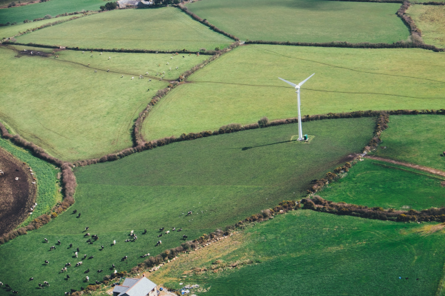 Luftaufnahme eines Windrades in einer grünen Wiese mit Bäumen, Häusern und Tieren in Irland.