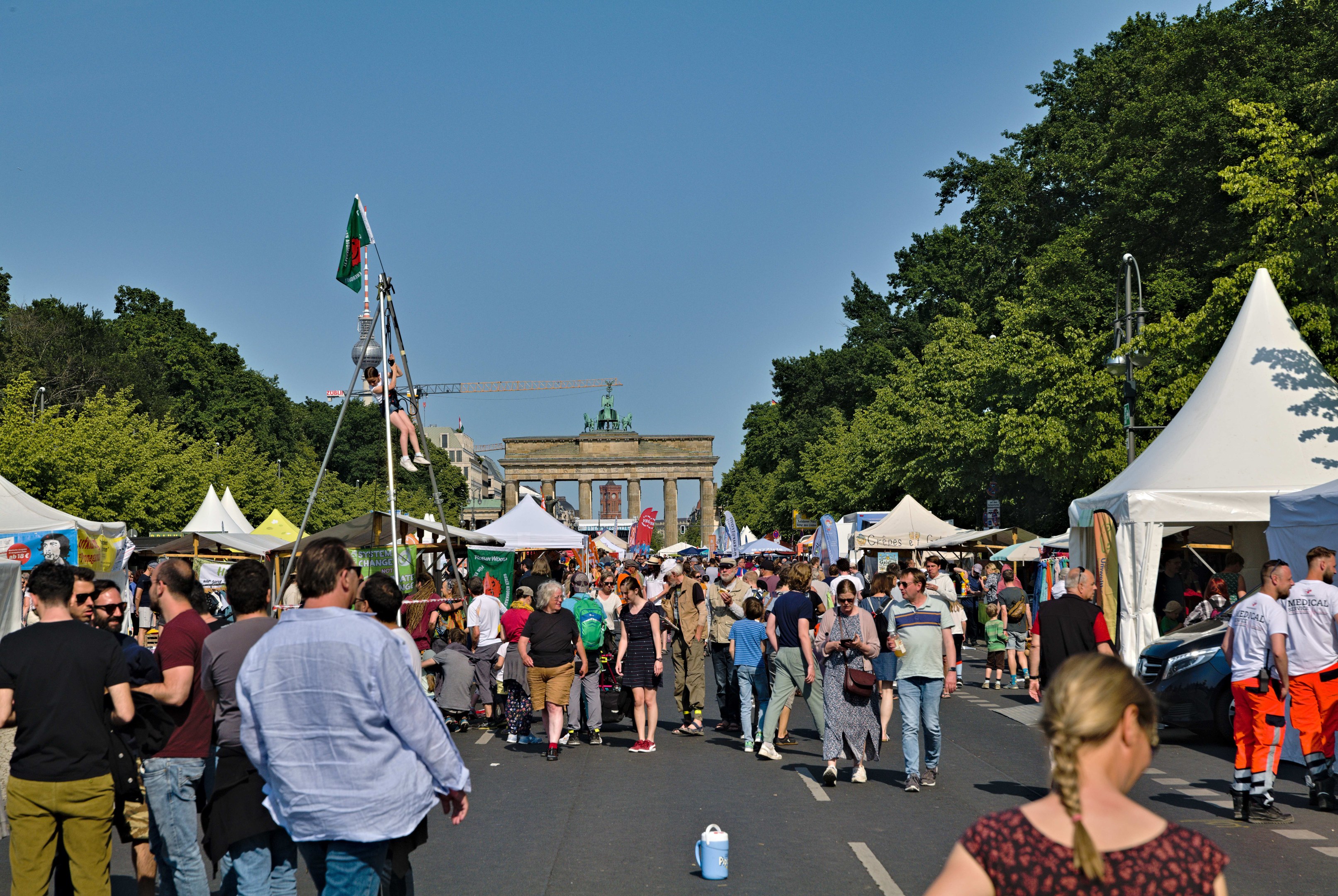 Eine Menge Menschen, die eine Straße entlanggehen, gesäumt von Zelten, Fahrzeugen und Bäumen, mit einem Bogen und einem klaren blauen Himmel im Hintergrund und Masten mit Flaggen auf der linken Seite, wahrscheinlich das Oktoberfest in München, Deutschland darstellend.