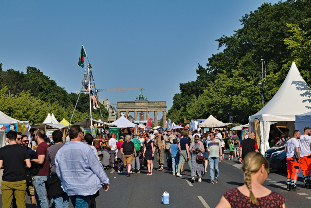 Eine Menge Menschen, die eine Straße entlanggehen, gesäumt von Zelten, Fahrzeugen und Bäumen, mit einem Bogen und einem klaren blauen Himmel im Hintergrund und Masten mit Flaggen auf der linken Seite, wahrscheinlich das Oktoberfest in München, Deutschland darstellend.