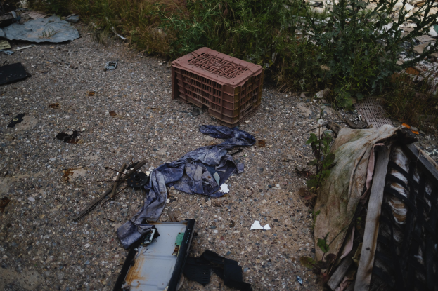 Ein Haufen Müll, einschließlich einer Plastikbox, Kleidung und anderen Gegenständen, der auf dem Boden neben einem Zaun verstreut liegt, mit Pflanzen und Gras im Hintergrund.
