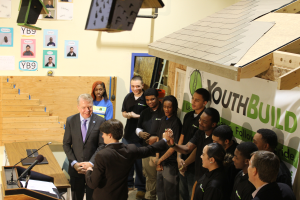 Eine Gruppe von Menschen steht um ein Rednerpult vor einer Menge, einige schütteln Hände, ein Tisch mit Mikrofonen und Papieren links, Plakate an der Wand im Hintergrund und Gegenstände auf dem Dach und der rechten Seite.