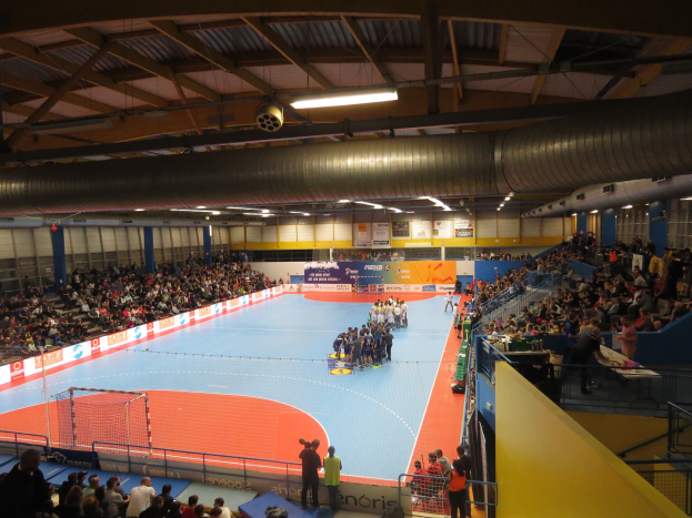 Große Halle mit Zuschauern bei einem Handballspiel, mit Sitzplätzen und Stehplätzen, einem Netz auf der linken Seite, Anzeigetafeln im Hintergrund und Beleuchtung und Rohren oben.