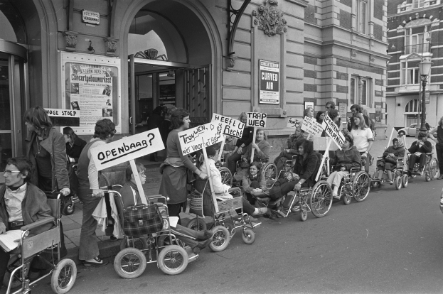 Eine Gruppe von Menschen im Rollstuhl mit Schildern am Straßenrand, im Hintergrund Gebäude und ein Laternenmast auf der rechten Seite, in Schwarz-Weiß.
