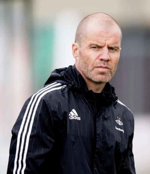 Ein kahlköpfiger Trainer in einer blauen Jacke mit weißen Streifen und einem weißen Kragen steht auf einem Fußballfeld mit verschränkten Händen und blickt ernsthaft nach vorne.