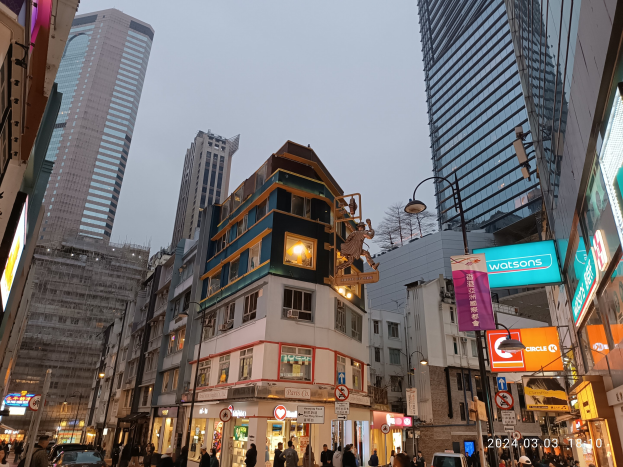 Eine belebte Straße mit hohen Gebäuden, fahrenden Fahrzeugen, Passanten, Straßenlaternen, Schildern und Baustellenaktivitäten im Vordergrund unter einem sichtbaren Himmel.