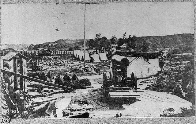 Schwarzes und weißes Foto einer Baustelle mit einer Maschine im Vordergrund, umgeben von Hütten, Bäumen, Hügeln und einem Himmel im Hintergrund, mit Text unten, der besagt: "Die Bauarbeiten an einer neuen Brücke in der Stadt San Francisco, Kalifornien."