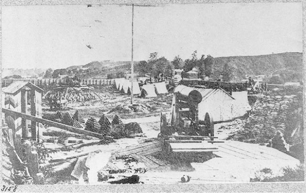 Schwarzes und weißes Foto einer Baustelle mit einer Maschine im Vordergrund, umgeben von Hütten, Bäumen, Hügeln und einem Himmel im Hintergrund, mit Text unten, der besagt: "Die Bauarbeiten an einer neuen Brücke in der Stadt San Francisco, Kalifornien."