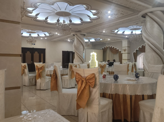 Großer Raum mit Tischen und Stühlen, die für eine Hochzeitsfeier arrangiert sind, Stühle mit Tüchern drapiert und mit dekorativen Accessoires versehen, Wände mit Türen und Vorhängen geschmückt, Decke mit Lichtern und Kronleuchtern dekoriert.