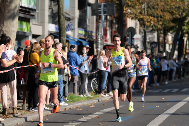 Gruppe von Läufern beim Marathon auf einer Stadtstraße, mit Zuschauern auf dem linken Gehweg, Bäumen und Gebäuden im Hintergrund unter einem klaren blauen Himmel.