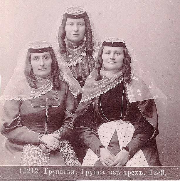 Drei Frauen in traditionellen russischen Hochzeitskleidern mit aufwendiger Stickerei und Perlen posieren vor einer weißen Wand in einem Schwarz-Weiß-Foto.