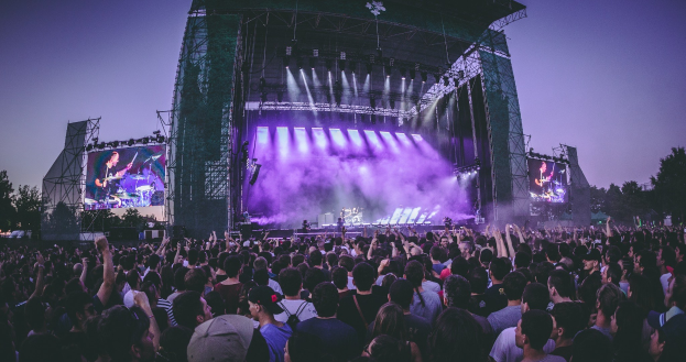 Eine große Menge versammelt sich auf einem Musikfestival mit einer Bühne, auf der Musiker spielen, beleuchtet von Lichtern und Bildschirmen, vor einer Kulisse aus Gebäude, Bäumen und einem klaren blauen Himmel.