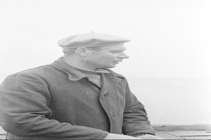 Ein altes Schwarz-Weiß-Foto eines Mannes in Jacke und Mütze, der nach rechts schaut und eine Uhr am Handgelenk trägt.