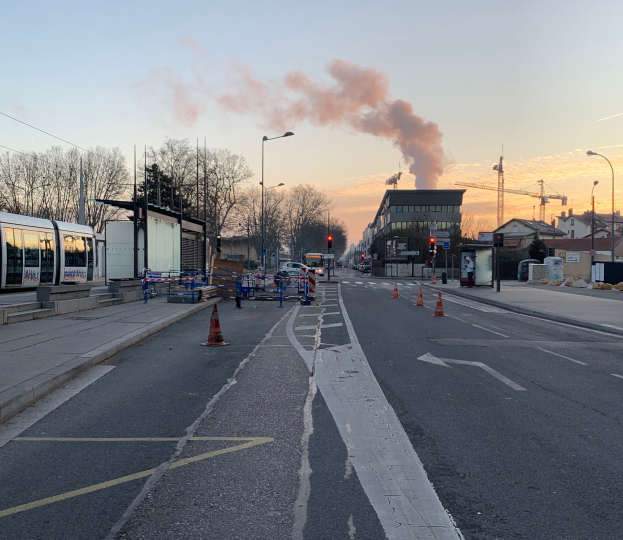 Eine Stadtstraße mit Absperrbaken, einem Bus, Laternenpfählen, Bäumen, Gebäuden, Kränen und Rauch, der aus einem fernen Gebäude aufsteigt, unter einem Himmel.