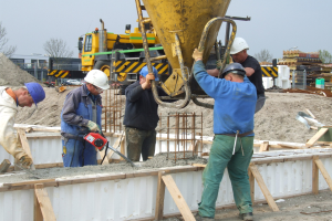 Gruppe von Männern mit Helmen bei der Arbeit auf einer Baustelle, einer Bedient ein Gerät, während die anderen Beton in einen Mischer gießen, mit Fahrzeugen, Bäumen, Gebäuden und einem klaren blauen Himmel im Hintergrund.