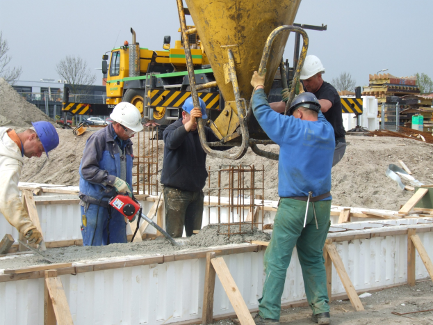 Gruppe von Männern mit Helmen bei der Arbeit auf einer Baustelle, einer Bedient ein Gerät, während die anderen Beton in einen Mischer gießen, mit Fahrzeugen, Bäumen, Gebäuden und einem klaren blauen Himmel im Hintergrund.