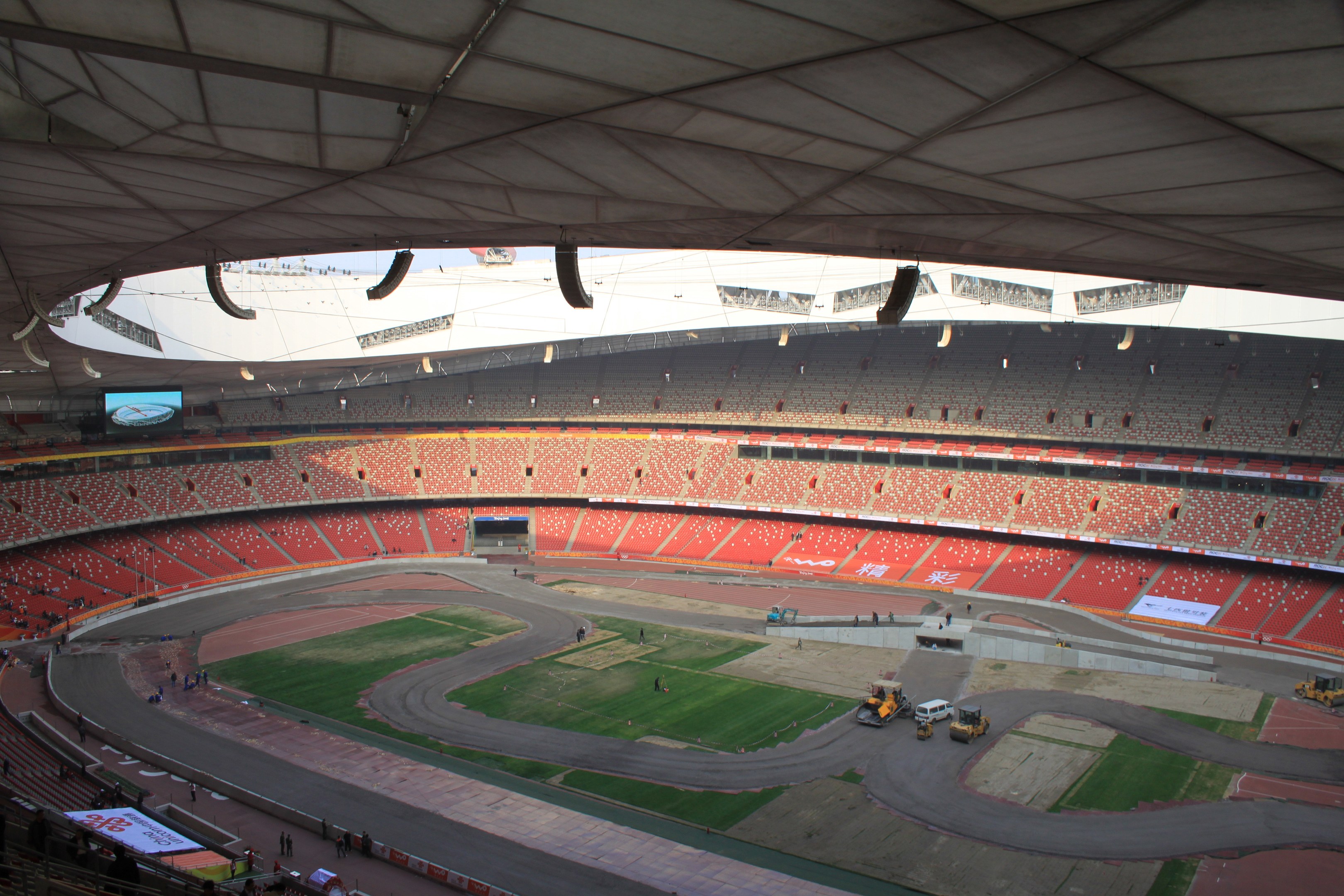 Großes Stadion voller Zuschauer, umgeben von Fahrzeugen auf einer Straße, Grasfläche, Texttafeln, Geländer, ein Bildschirm und Überdachung.