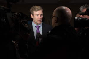 Ein Mann im Anzug und Krawatte, der eine Brille trägt, spricht zu Reportern mit zwei Kameramännern auf jeder Seite und einem Mikrofon vor ihm.