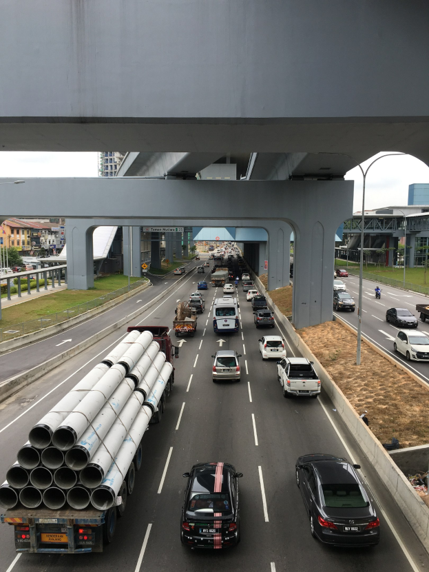Eine befahrene Autobahn mit zahlreichen Fahrzeugen, eine Brücke darüber, Straßenlaternen, Gras, Gebäude, Bäume und einen klaren Himmel im Hintergrund.