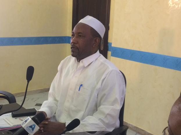 Ein Mann in weißem Anzug und Hut sitzt an einem Tisch mit Mikrofonen, spricht, während Papiere und Gegenstände auf dem Tisch zu sehen sind, mit einem anderen Menschen mit Brille rechts von ihm, in einem Raum mit einer Wand und einer Tür dahinter.