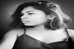 Eine selbstbewusste Frau in einem schwarzen Kleid posiert in einem Schwarz-Weiß-Foto, ihr glatter Bob und das dezente Make-up unterstreichen ihre selbstbewusste Haltung vor einer neutralen Wand.