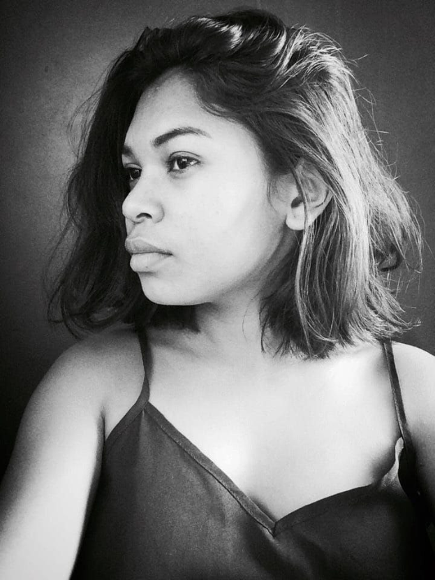 Eine selbstbewusste Frau in einem schwarzen Kleid posiert in einem Schwarz-Weiß-Foto, ihr glatter Bob und das dezente Make-up unterstreichen ihre selbstbewusste Haltung vor einer neutralen Wand.