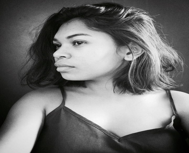 Eine selbstbewusste Frau in einem schwarzen Kleid posiert in einem Schwarz-Weiß-Foto, ihr glatter Bob und das dezente Make-up unterstreichen ihre selbstbewusste Haltung vor einer neutralen Wand.