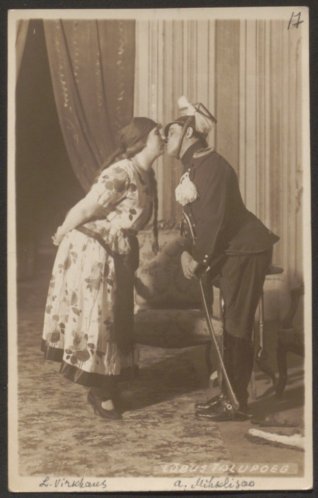 Schwarzes und weißes Foto eines Mannes und einer Frau, die sich küssen, wobei der Mann einen Stock hält und die Frau neben ihm steht, ein Stuhl und ein Vorhang im Hintergrund und Text unten.