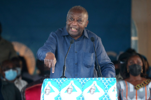 Ein Mann in einem blauen Hemd spricht an einem Podium mit zwei Mikrofonen vor einem sitzenden Publikum, einige tragen Masken, mit einer Wand im Hintergrund.